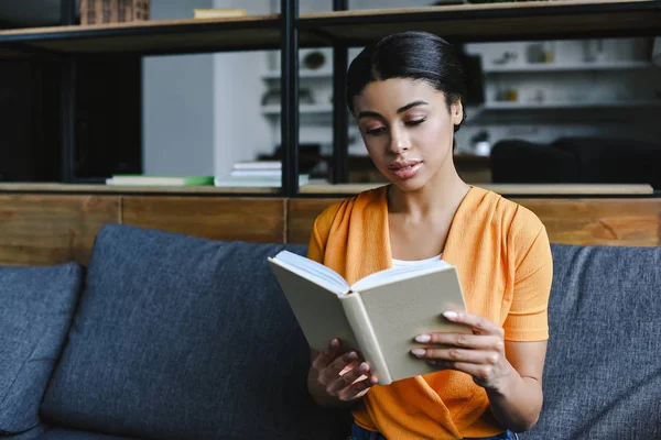 Bela menina de raça mista em livro de leitura camisa laranja no sofá na sala de estar — Fotografia de Stock