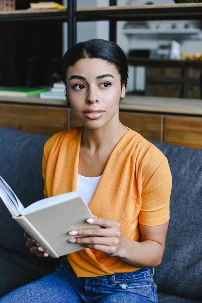 Hermosa chica de raza mixta en camisa naranja sosteniendo libro en la sala de estar y mirando hacia otro lado - foto de stock