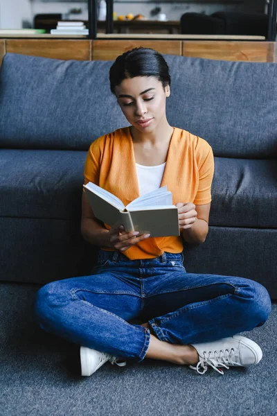 Belle fille de race mixte en chemise orange livre de lecture sur le sol dans le salon — Photo de stock