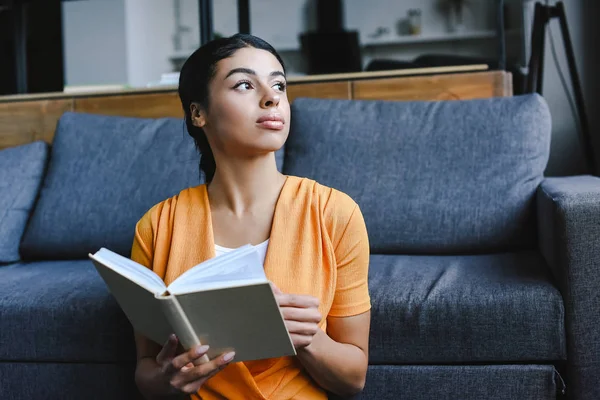 Bela menina de raça mista na camisa laranja segurando livro na sala de estar e olhando para longe — Fotografia de Stock