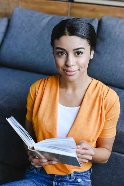 Beautiful mixed race girl in orange shirt holding book in living room and looking at camera — Stock Photo