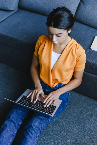High angle view of beautiful mixed race girl in orange shirt sitting on floor and using laptop in living room — Stock Photo
