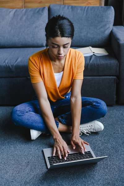 Schönes gemischtes Rennmädchen in orangefarbenem Hemd sitzt auf dem Boden und benutzt Laptop im Wohnzimmer — Stockfoto