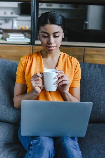 Hermosa chica de raza mixta en camisa naranja sosteniendo taza de café y el uso de ordenador portátil en la sala de estar - foto de stock