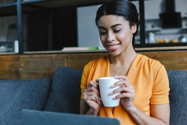 Sorridente bella ragazza razza mista in camicia arancione utilizzando il computer portatile e tenendo il tè tazza in soggiorno — Foto stock