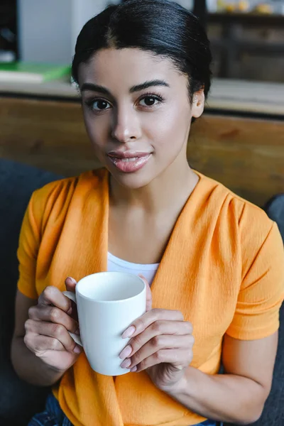 Portrait de belle fille de race mixte en chemise orange tenant tasse de thé et regardant la caméra à la maison — Photo de stock