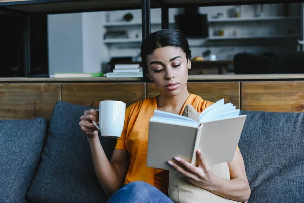 Atractiva chica de raza mixta en camisa naranja sosteniendo taza de té y libro de lectura en el sofá en casa - foto de stock