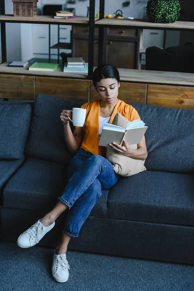 Vista ad alto angolo di bella ragazza di razza mista in camicia arancione con in mano tazza di tè e libro di lettura sul divano a casa — Foto stock