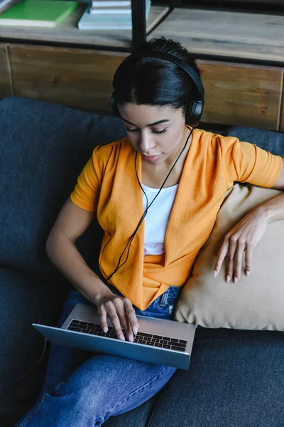 Vista de ángulo alto de hermosa chica de raza mixta en camisa naranja escuchar música y el uso de ordenador portátil en la sala de estar - foto de stock