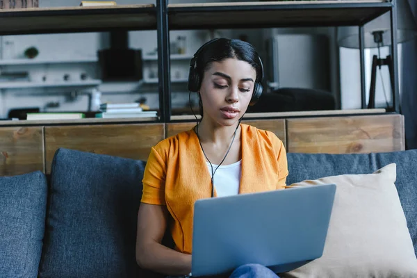 Schöne gemischte Rasse Mädchen in orangefarbenem Hemd Musik hören und mit Laptop im Wohnzimmer — Stockfoto