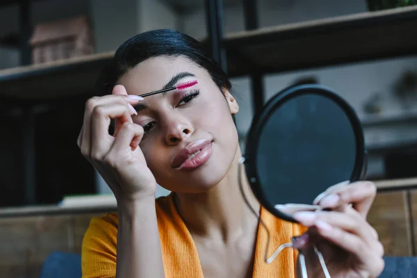 Retrato de hermosa chica de raza mixta en camisa naranja aplicando rímel en casa - foto de stock