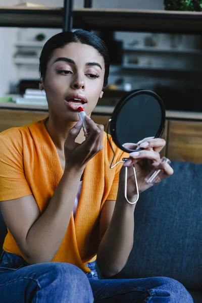 Beautiful mixed race girl in orange shirt applying red lipstick at home — Stock Photo