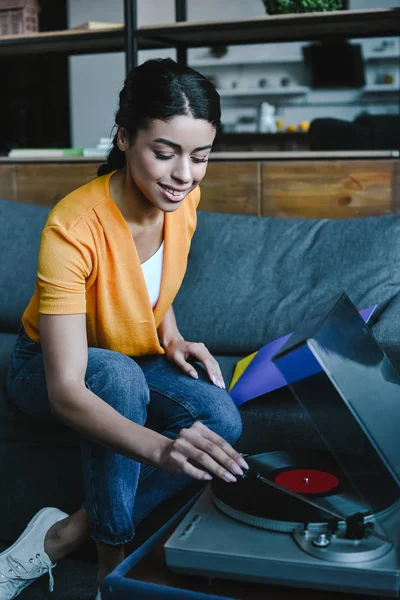 Sorridente bella ragazza di razza mista in camicia arancione girando sul grammofono con vinile retrò a casa — Foto stock