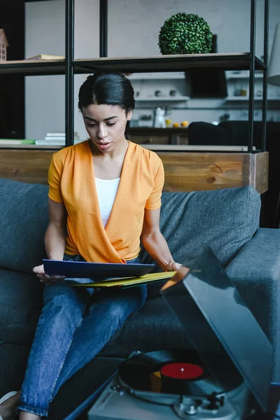 Atractiva chica de raza mixta en camisa naranja elegir vinilo en el sofá en la sala de estar - foto de stock