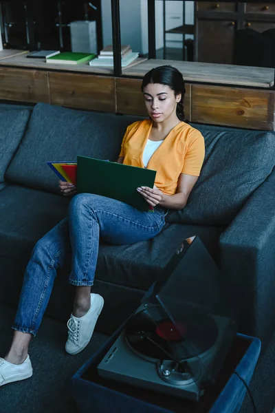 Hermosa chica de raza mixta en camisa naranja elegir vinilo en el sofá en la sala de estar - foto de stock