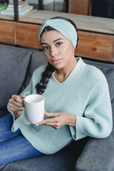 Attractive mixed race girl in turquoise sweater and headband sitting on sofa and holding cup of tea in living room — Stock Photo