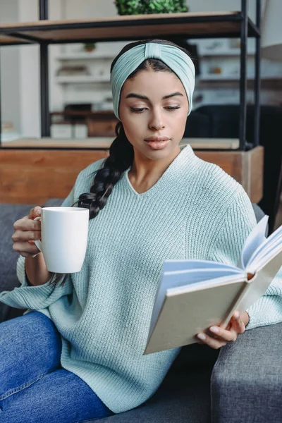 Atractiva chica de raza mixta en suéter turquesa y diadema sentado en el sofá y la celebración de la taza de té y libro de lectura en la sala de estar - foto de stock