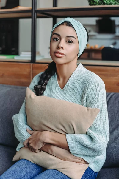 Atractiva chica de raza mixta en suéter turquesa y diadema abrazando almohada en el sofá en la sala de estar - foto de stock