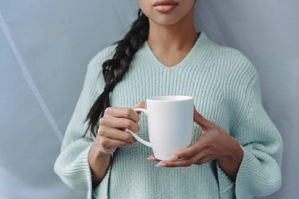 Imagen recortada de chica de raza mixta en suéter turquesa sosteniendo taza de té en casa - foto de stock