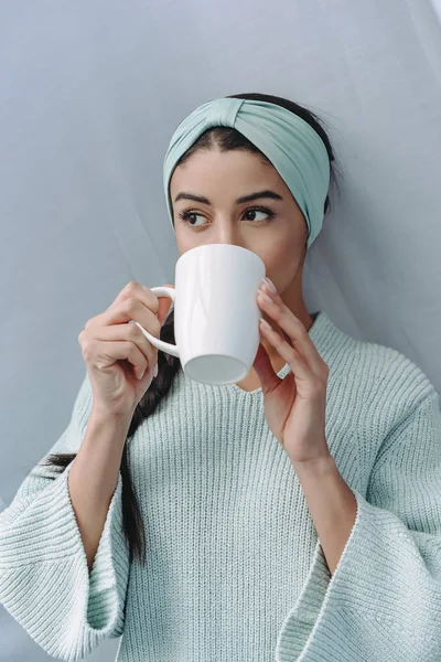 Atractiva chica de raza mixta en suéter turquesa y diadema bebiendo té en casa - foto de stock