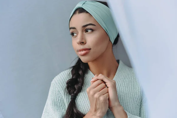 Atractiva chica de raza mixta en suéter turquesa y diadema mirando hacia fuera en casa - foto de stock