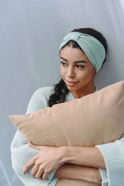 Hermosa chica de raza mixta en suéter turquesa y diadema abrazando almohada y mirando hacia otro lado - foto de stock