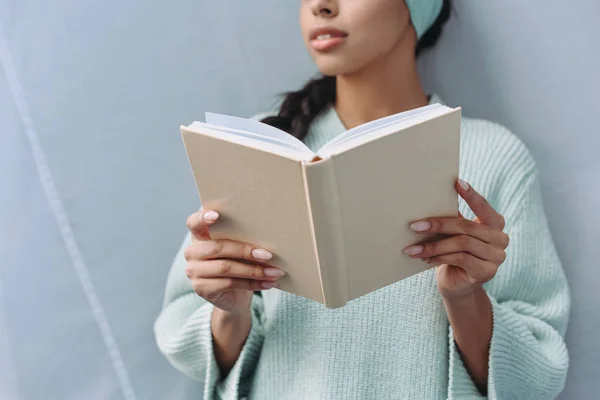 Image recadrée de fille de race mixte en pull turquoise et serre-tête tenant livre à la maison — Photo de stock