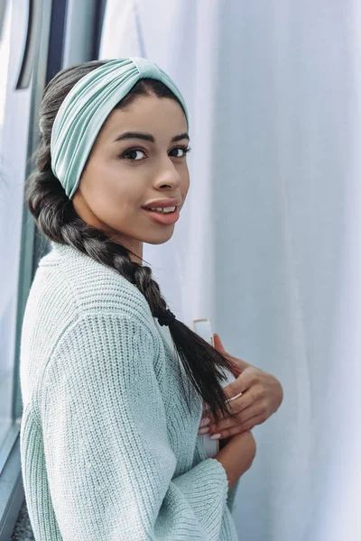 Hermosa chica de raza mixta en suéter turquesa y diadema sosteniendo libro y mirando a la cámara en casa - foto de stock