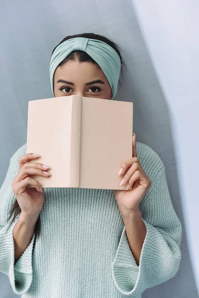Bela menina de raça mista em camisola turquesa e headband olhando para fora do livro em casa — Fotografia de Stock