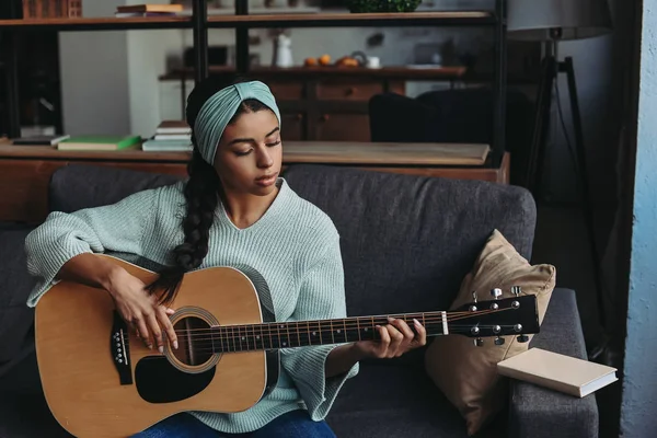 Hermosa chica de raza mixta en suéter turquesa y diadema tocando la guitarra acústica en el sofá en casa - foto de stock