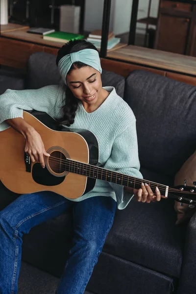 Blick aus der Vogelperspektive auf das schöne Mixed Race Girl in türkisfarbenem Pullover und Stirnband, das zu Hause auf dem Sofa Akustikgitarre spielt — Stockfoto