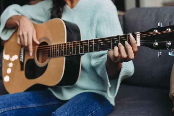Immagine ritagliata di ragazza razza mista in maglione turchese suonare la chitarra acustica sul divano a casa — Foto stock