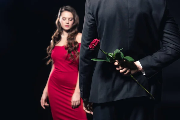 Selective focus of man hiding rose behind back with woman in red dress on background isolated on black — Stock Photo