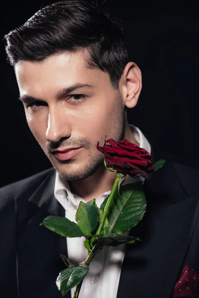 Портрет красивого чоловіка в костюмі, який тримає червону троянду ізольовано на чорному — стокове фото