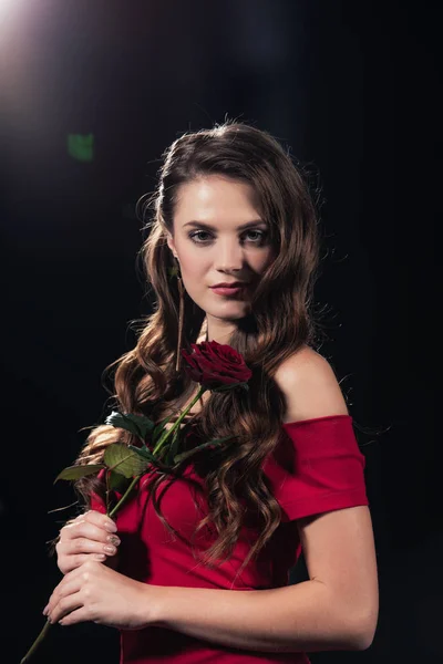 Beautiful woman in red dress looking at camera and holding rose on black background — Stock Photo