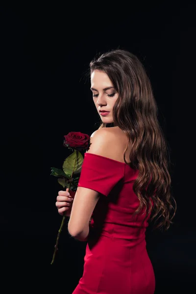 Hermosa mujer en vestido rojo con los ojos cerrados sosteniendo rosa aislado en negro - foto de stock