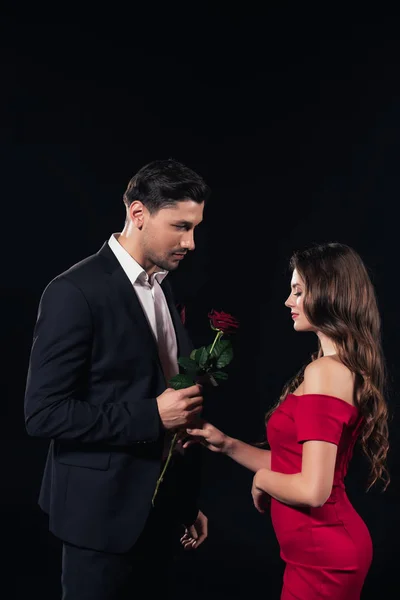 Hombre guapo presentando rosa roja a mujer en vestido rojo aislado en negro - foto de stock