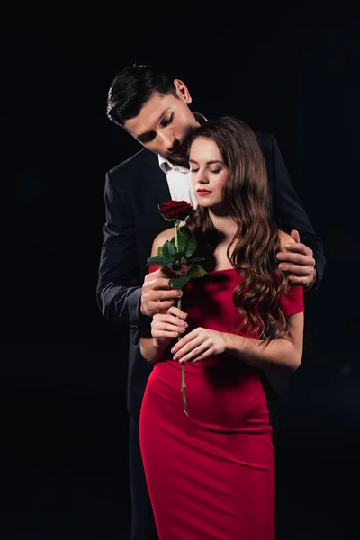 Homem bonito abraçando mulher bonita em vestido vermelho segurando rosa isolada no preto — Fotografia de Stock