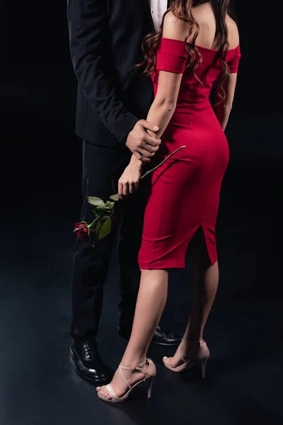 Vista recortada del hombre abrazando a la mujer en vestido rojo sosteniendo rosa aislado en negro - foto de stock