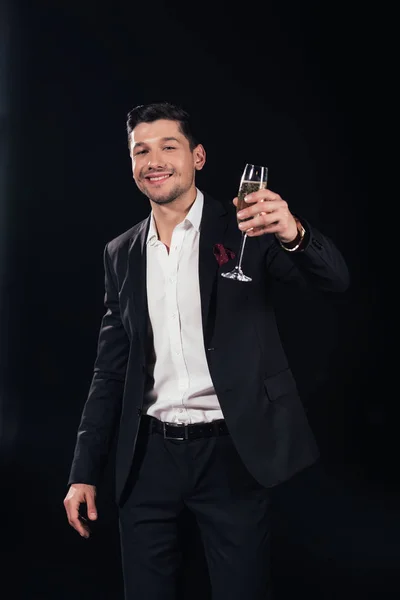 Souriant bel homme regardant la caméra et soulevant verre de champagne isolé sur noir — Photo de stock