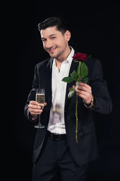 Hombre guapo sonriente mirando a la cámara y sosteniendo una copa de champán con rosa roja aislada en negro - foto de stock