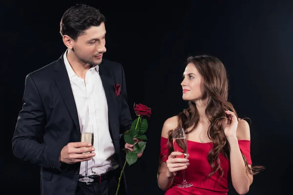 Hermosa pareja mirándose, sosteniendo rosa roja y copas de champán aisladas en negro - foto de stock