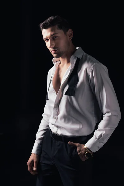 Hombre guapo en camisa blanca y mano en bolsillo mirando a la cámara mientras posando aislado en negro - foto de stock