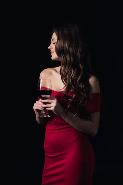 Hermosa mujer en vestido rojo sosteniendo copa de vino tinto y mirando hacia otro lado aislado en negro - foto de stock