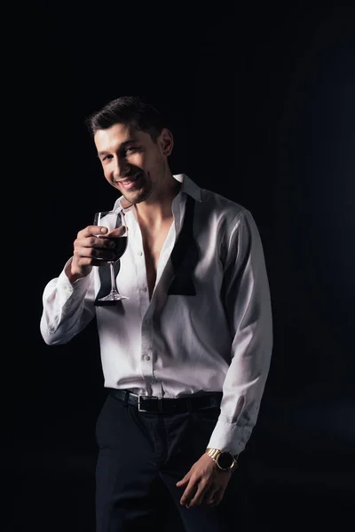 Homem sorridente em camisa branca olhando para câmera e segurando vidro de vinho tinto isolado em preto — Fotografia de Stock