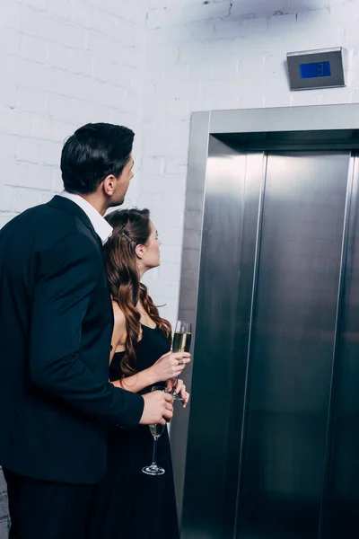 Couple in formal wear holding champagne glasses and waiting for elevator — Stock Photo