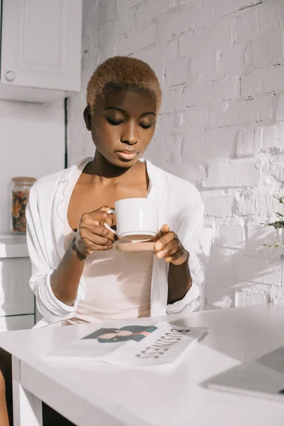 Atractiva mujer afroamericana sosteniendo taza en cocina blanca - foto de stock
