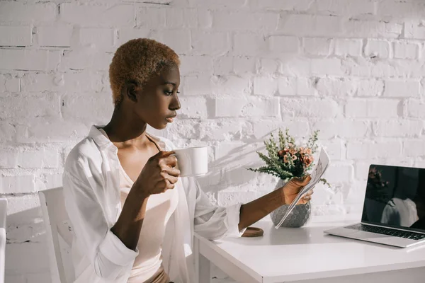 Mujer afroamericana leyendo periódico de negocios y sosteniendo taza de café - foto de stock