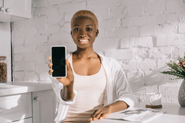 Femme américaine africaine joyeuse montrant smartphone avec écran blanc dans la cuisine blanche — Photo de stock