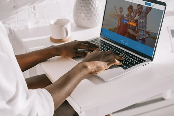 Vista recortada de la mujer afroamericana escribiendo en el ordenador portátil con couchsurfing sitio web - foto de stock